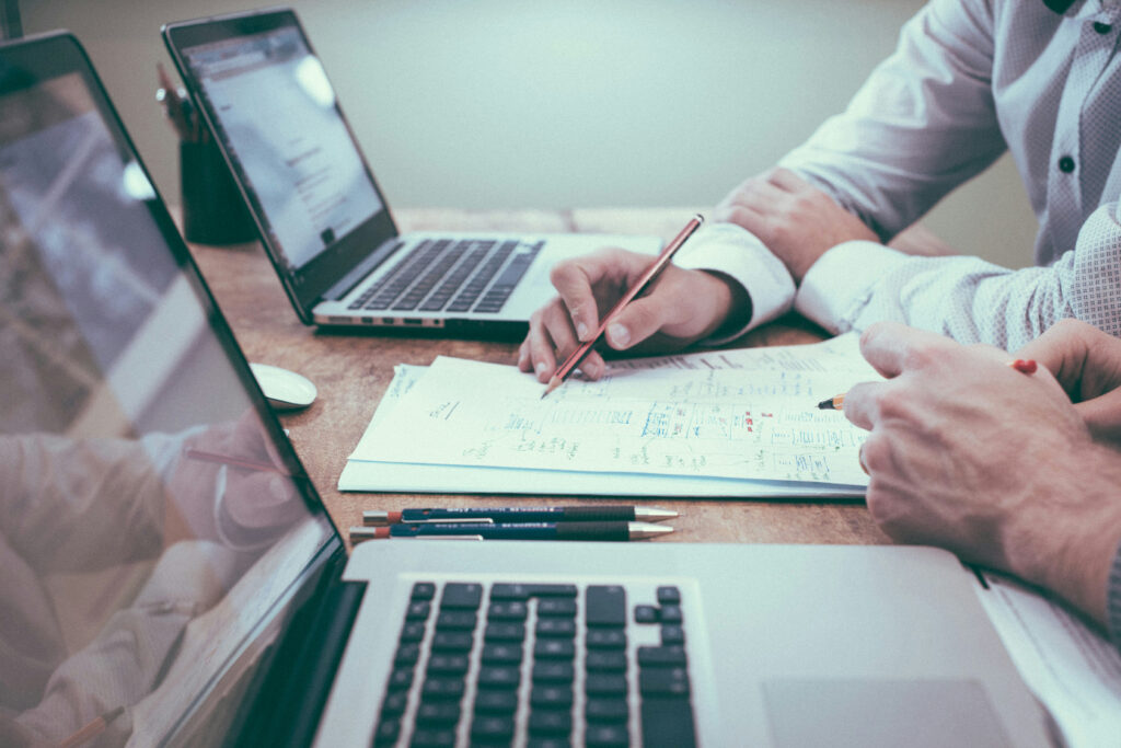 Two people in formal attire are seated at a wooden table with laptops open, discussing and making notes on documents. One person is pointing at the paper with a pencil, sharing tips on optimizing their WordPress website. Pens are placed nearby, ready for action.