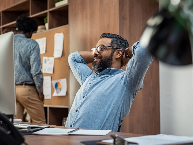 A man with glasses and a beard is sitting at a desk, smiling confidently with his hands behind his head. Papers and charts illustrate strategies for managed WordPress hosting on a board behind him, while another person studies the board intently.