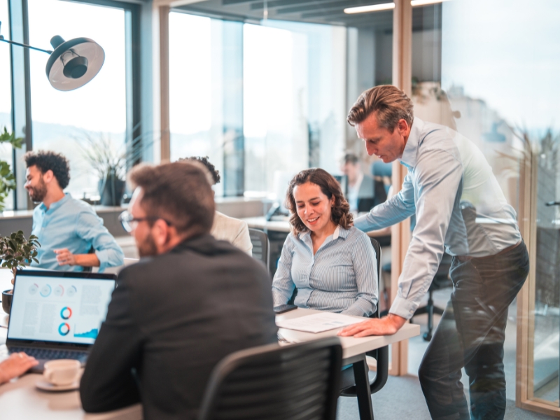 In a modern office setting, a group collaborates around a table. One person leans over to assist a seated colleague, who is smiling. A laptop displays charts, possibly related to WordPress maintenance, while large windows flood the space with natural light.