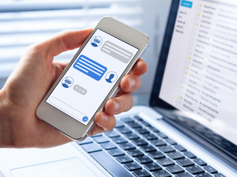 A person holds a smartphone displaying a chat screen with blue and gray message bubbles, while an open laptop in the background shows an email inbox alongside WordPress add-ons. The setting suggests seamless communication and multitasking.