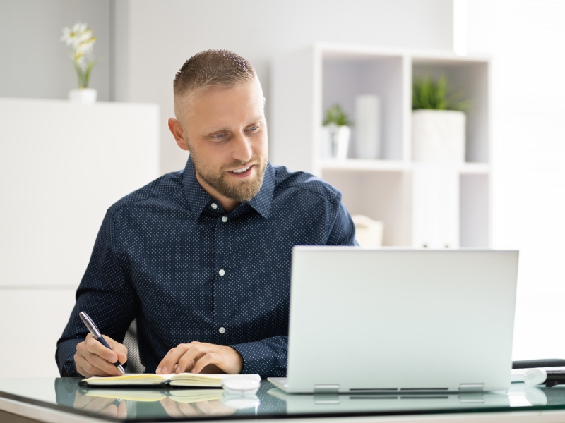 A man sits at a desk, intently studying WordPress Add-ons on his laptop while scribbling notes in a notebook. He wears a dark shirt and has short hair and a beard. In the background, shelves adorned with plants and office items create a serene workspace ambiance.