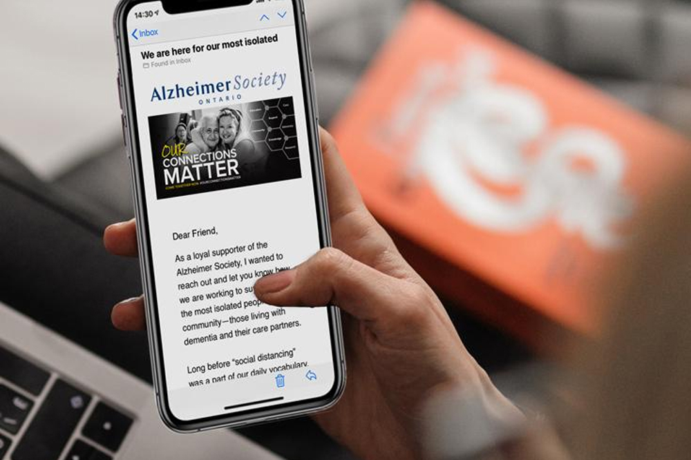 A person holding a smartphone displaying an email from Alzheimer Society Ontario with the subject "We are here for our most isolated." In the background, there’s a blurred orange object and part of a laptop used for WordPress maintenance.