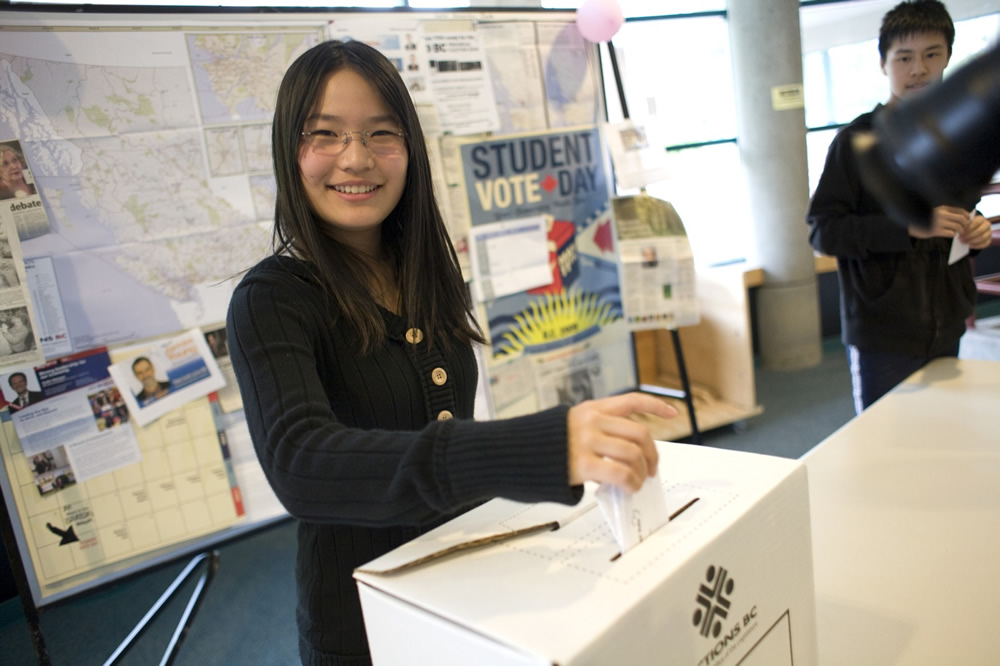 A young woman smiles as she places a ballot into a white voting box. It's "Student Vote Day," with maps and posters decorating the board behind her, reminiscent of the organization needed for WordPress hosting. Nearby, another person stands slightly out of focus.