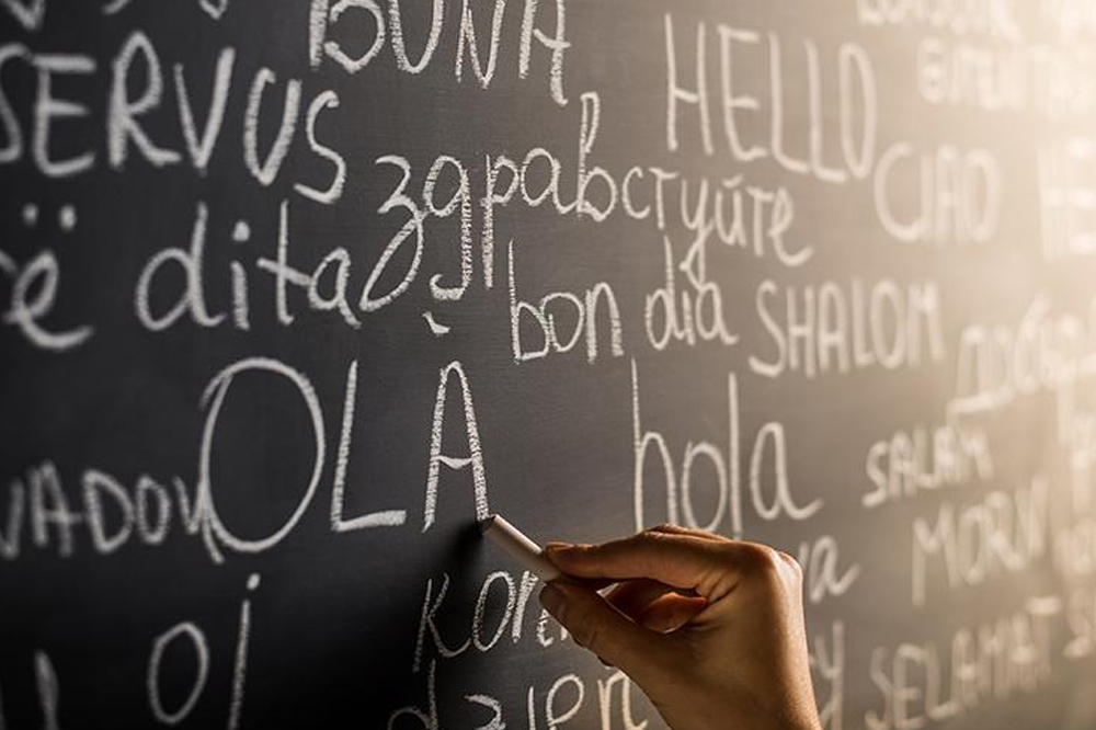 A person writes with white chalk on a blackboard filled with the word "hello" in various languages, including "OLA," "buna," "bonjour," and "hola." The board, angled slightly to capture part of the writer's hand, serves as an artistic metaphor for WordPress Hosting's global reach.