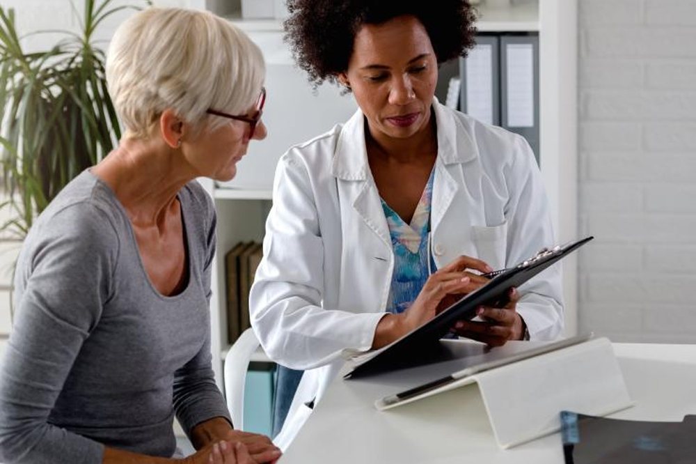 A doctor in a white coat discusses a medical chart with an older woman in an office. While showing information on a clipboard, he compares it to how WordPress Maintenance keeps sites healthy. The woman listens attentively, surrounded by the calming presence of a plant in the background.